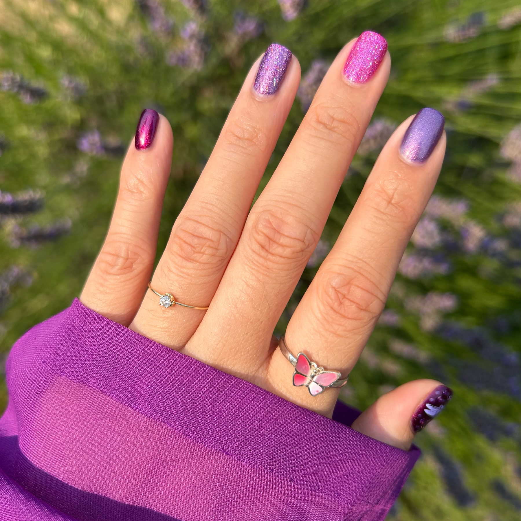 A close-up of a hand with manicured nails in purple and lavender shades, wearing a silver ring with a butterfly design and a small diamond, set against a blurred background of green grass and purple lavender flowers.
