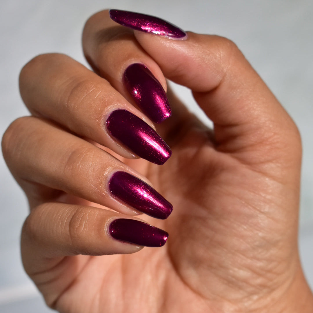 A close-up of a hand with long, almond-shaped nails painted in a metallic deep magenta-purple polish, set against a soft light gray background.