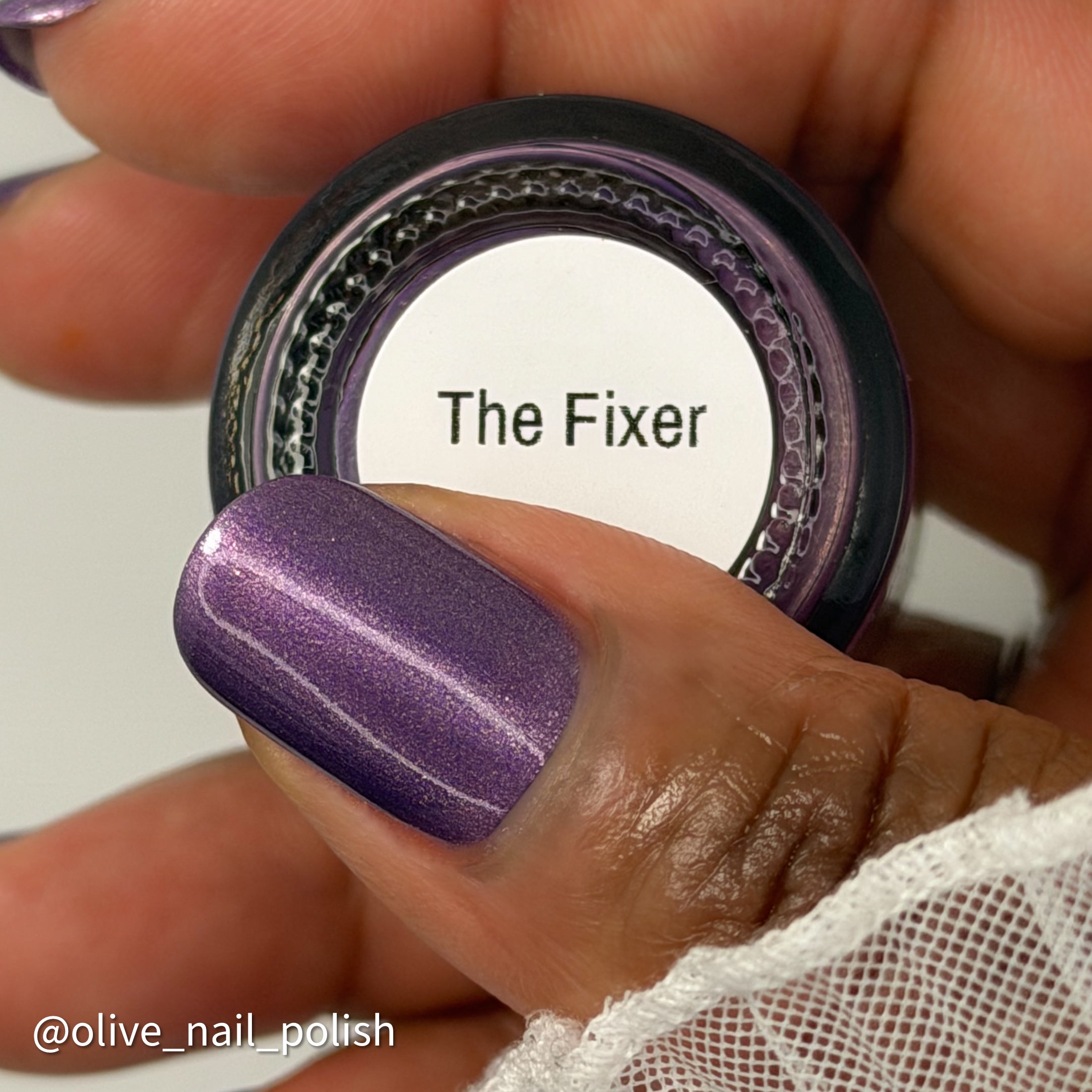 A close-up of a hand holding a small bottle of nail polish with the label reading 'The Fixer.' The visible nail polish color is a shimmery metallic purple, and the hand is wearing the polish on a fingernail.
