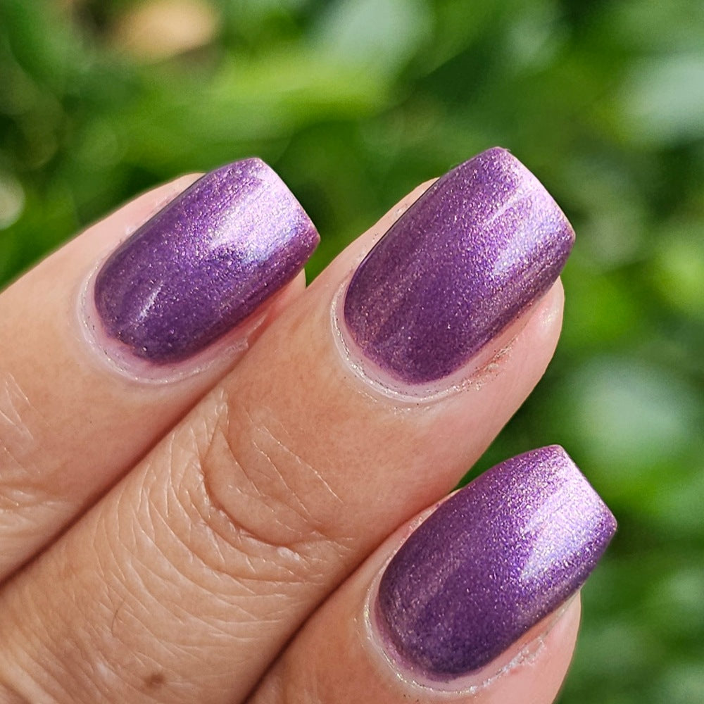 A close-up of a hand with manicured nails painted in a shimmering metallic purple polish, set against a blurred green outdoor background.