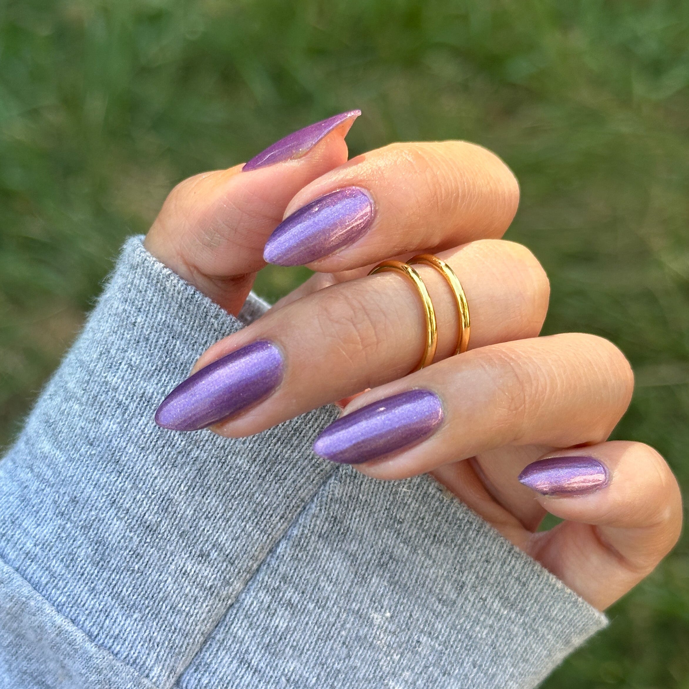 A close-up of a hand with long, almond-shaped nails painted in a shimmering metallic lavender-purple polish, wearing two thin gold rings, with a blurred green outdoor background.