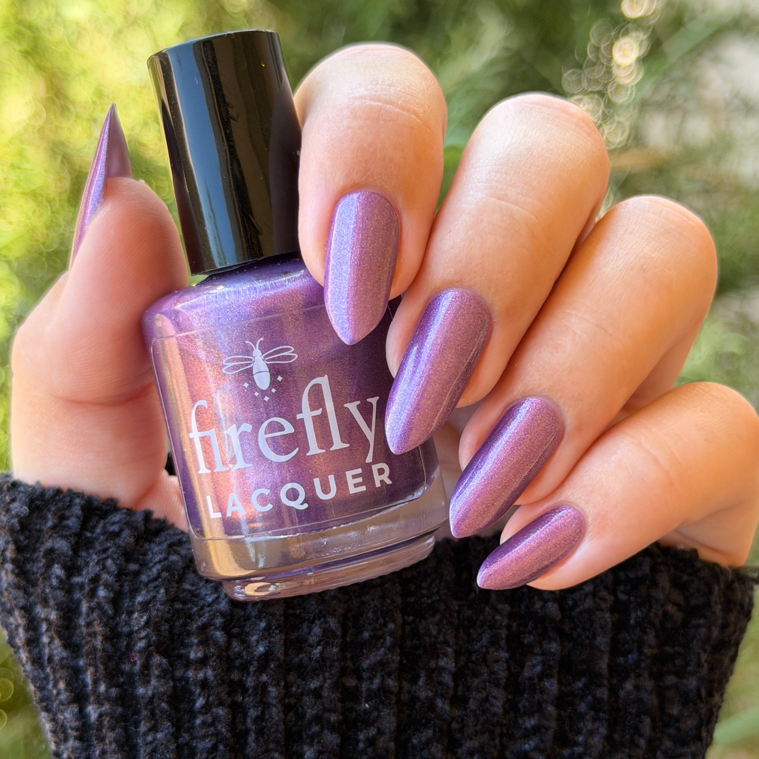 A hand with lavender-purple manicured nails holds a bottle of Firefly Lacquer nail polish in a matching shimmering purple shade, set against a blurred green outdoor background.