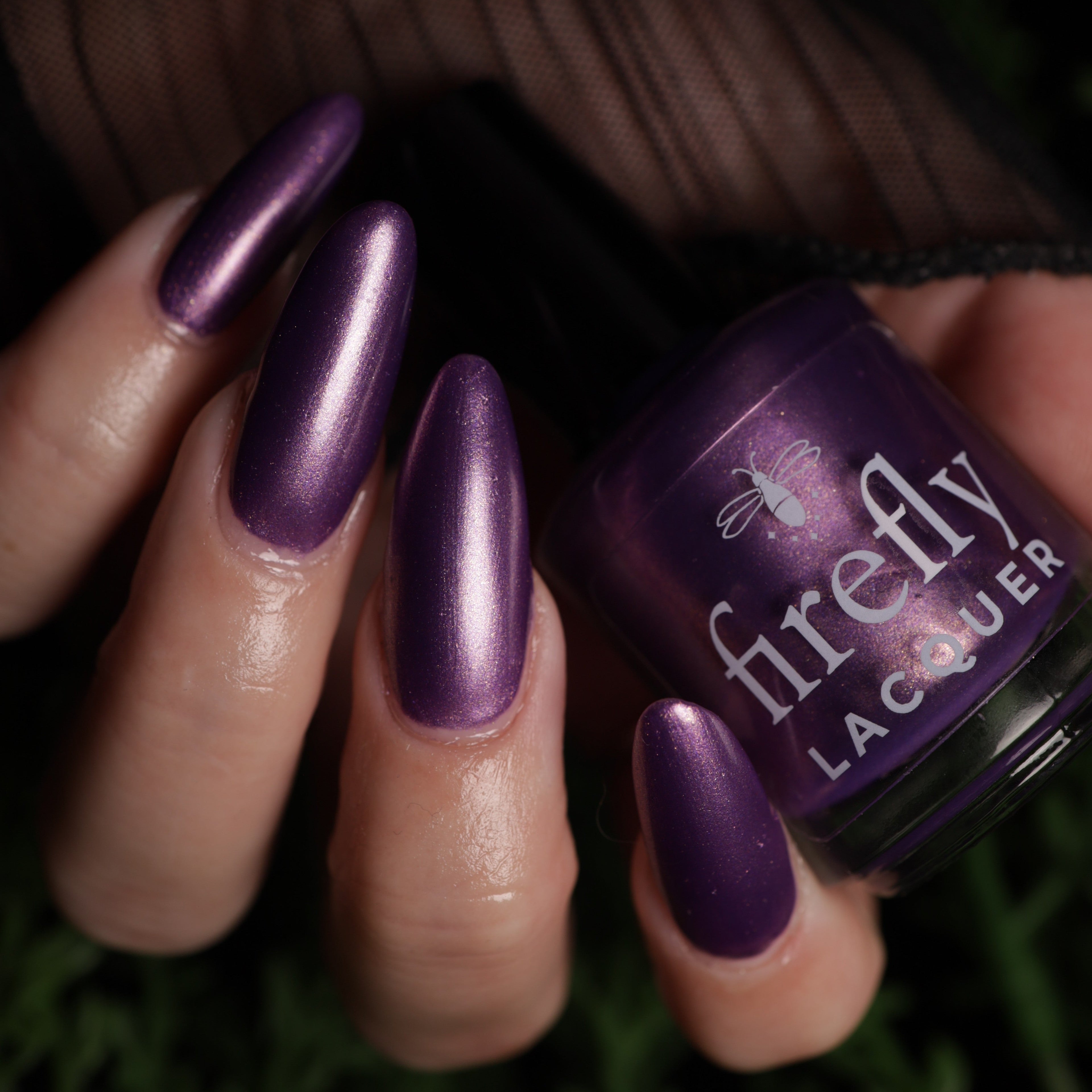 A close-up of a hand with long, glossy metallic purple nails holding a bottle of Firefly Lacquer in a matching deep purple shade.
