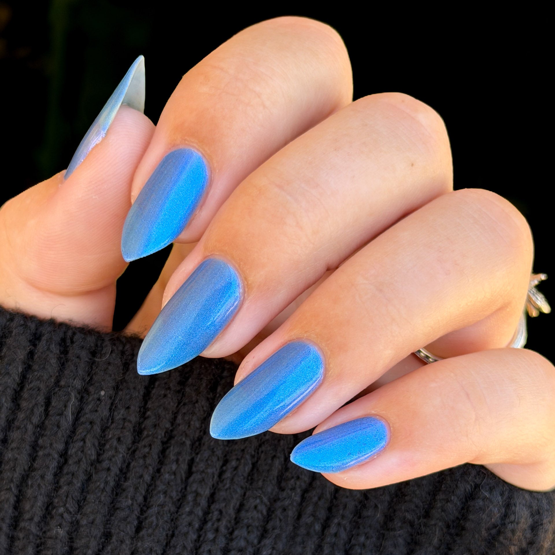 A close-up of a hand with long, almond-shaped nails painted in a vibrant blue ombre gradient, wearing a black ribbed sweater against a dark background.