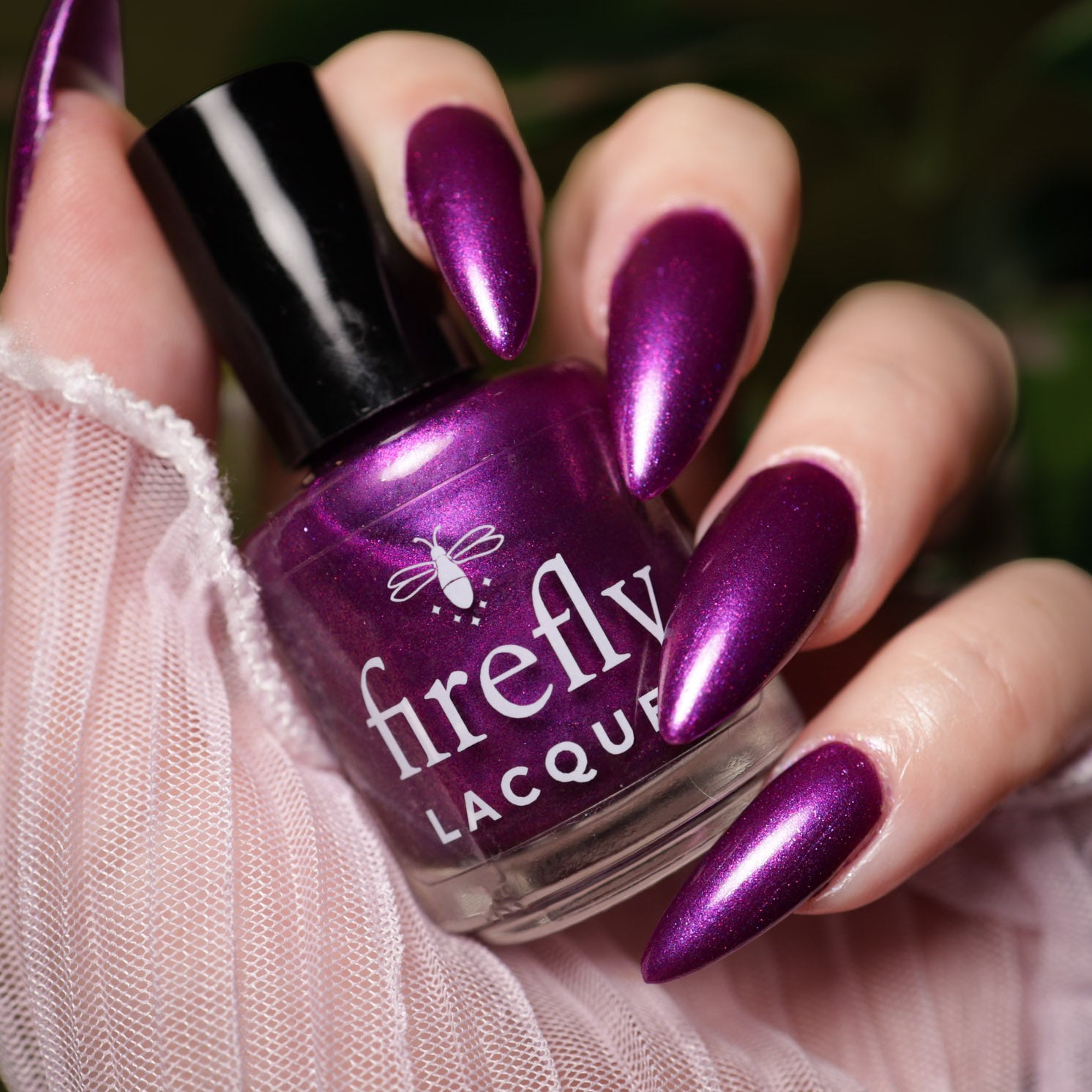 A close-up of a hand with long, metallic purple manicured nails holding a bottle of Firefly Lacquer nail polish in a matching deep shimmering purple shade.