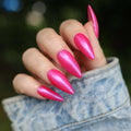 Hand with pink stiletto nails wearing a denim jacket against a blurred green background