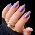 A close-up of a hand with long, almond-shaped nails painted in a shimmering metallic lavender-purple shade, wearing a dark knit sleeve and a silver ring against a black background.