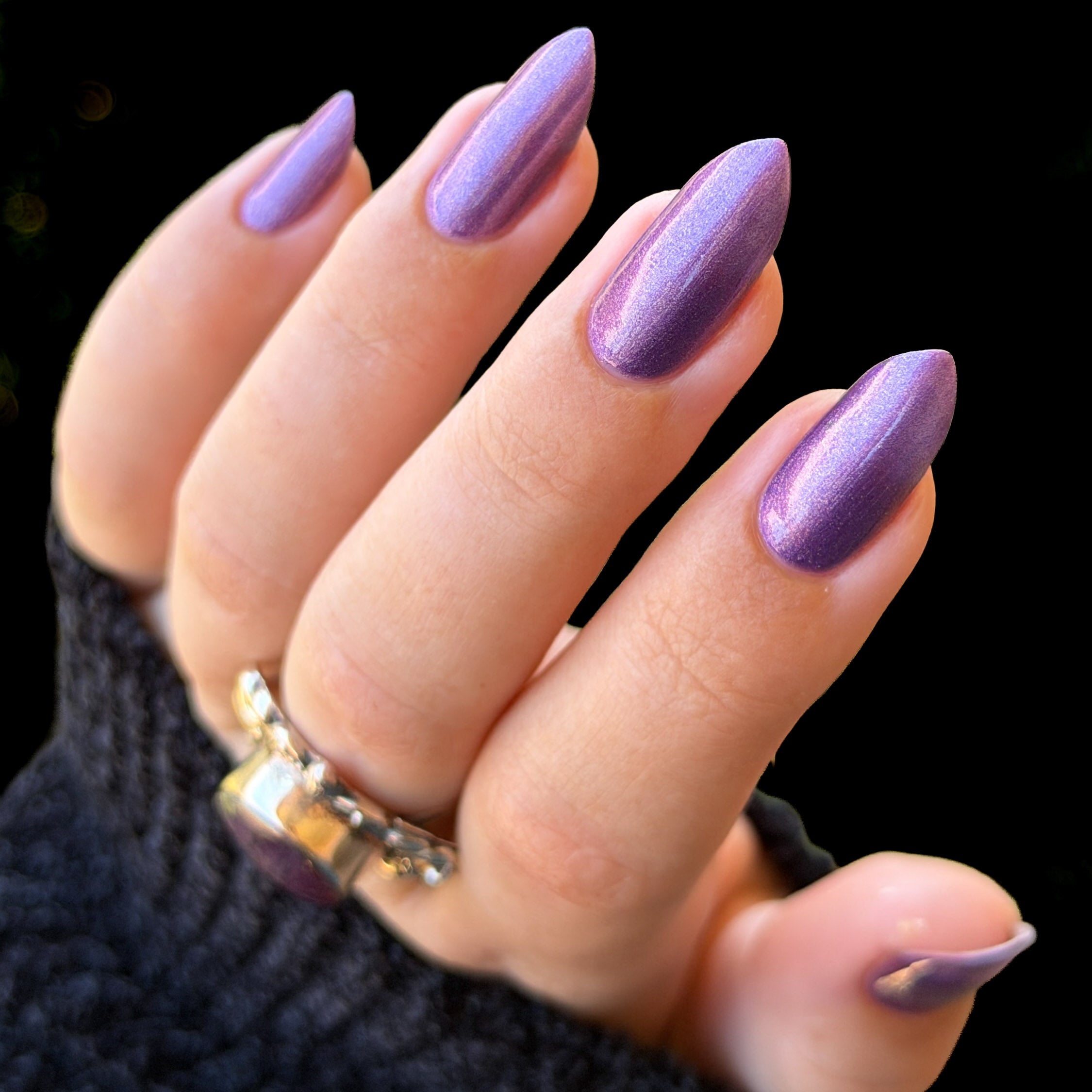 A close-up of a hand with long, almond-shaped nails painted in a shimmering metallic lavender-purple shade, wearing a dark knit sleeve and a silver ring against a black background.
