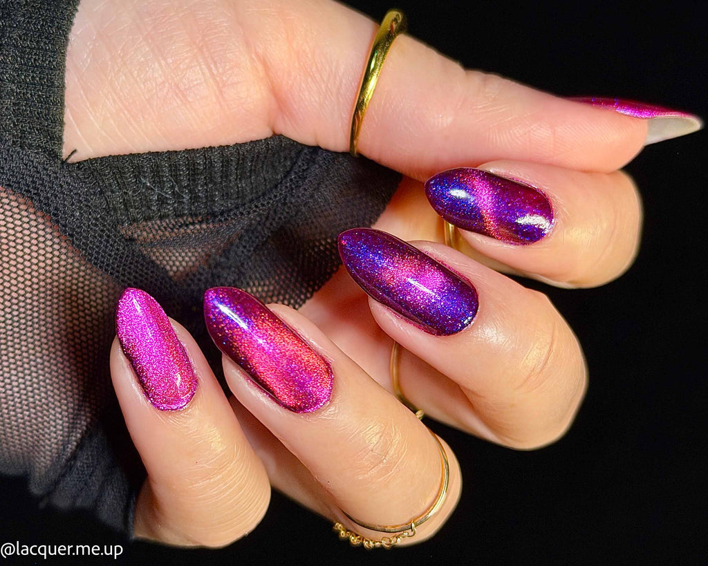 A close-up of a hand with long almond-shaped nails painted in a metallic magenta and purple iridescent nail polish, wearing gold rings against a black background.
