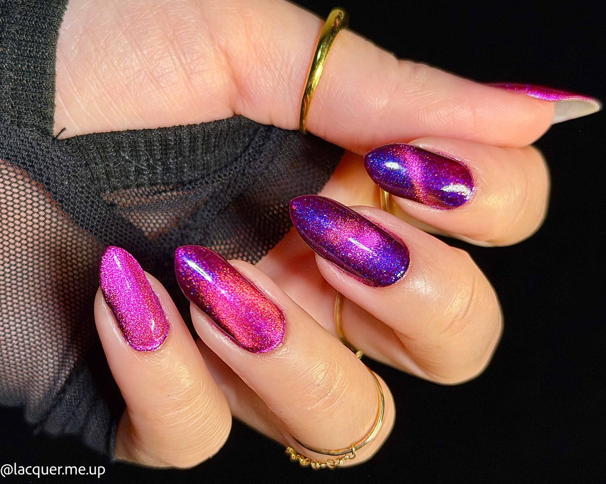 A close-up of a hand with long almond-shaped nails painted in a metallic magenta and purple iridescent nail polish, wearing gold rings against a black background.