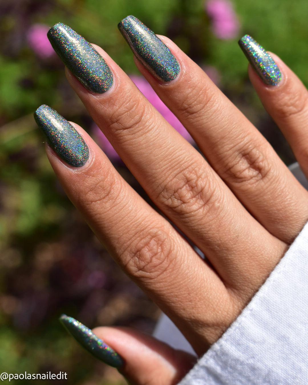 A close-up of a hand with long almond-shaped nails painted in a dark teal glitter nail polish with iridescent holographic shimmer, set against a blurred outdoor background with green foliage and pink flowers.
