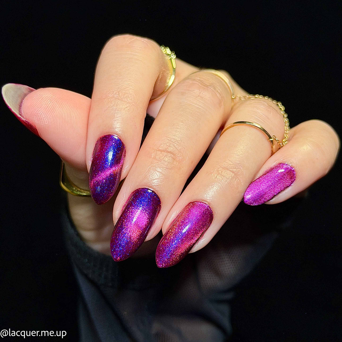 A close-up of a hand showcasing almond-shaped nails painted with a shimmering purple and pink cat-eye magnetic nail polish. The hand is adorned with several gold rings against a solid black background.