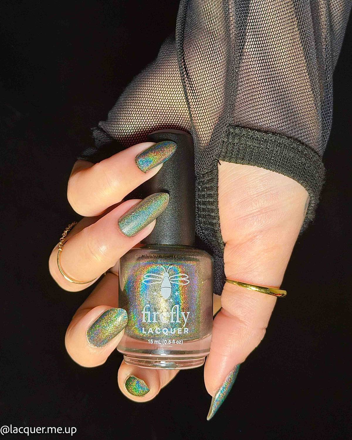 A hand holding a bottle of Firefly Lacquer nail polish against a black background. The nails are painted with a shimmering, iridescent green and gold holographic polish, and the hand is wearing sheer black mesh sleeves and gold rings.