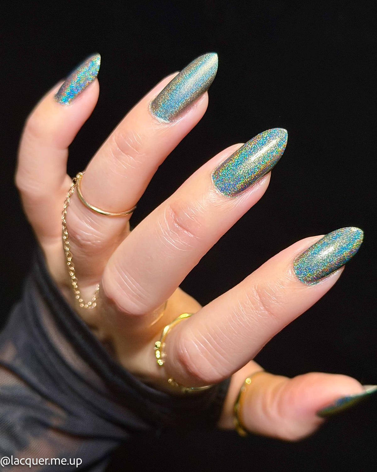 A close-up of a hand with long, almond-shaped nails painted in a shimmering teal-blue nail polish with gold glitter, wearing several gold rings against a black background.