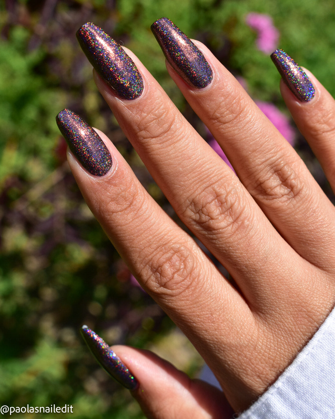 A close-up of a hand displaying long, almond-shaped nails painted with a dark, shimmery holographic glitter nail polish. The background is a soft-focus outdoor garden with green foliage and a hint of pink flowers.