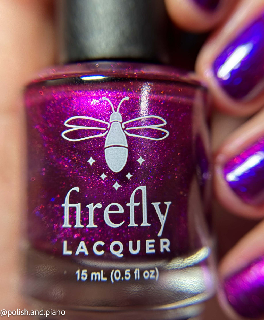 A close-up of a Firefly Lacquer nail polish bottle filled with a sparkling magenta-purple polish, held by a hand with matching painted fingernails.