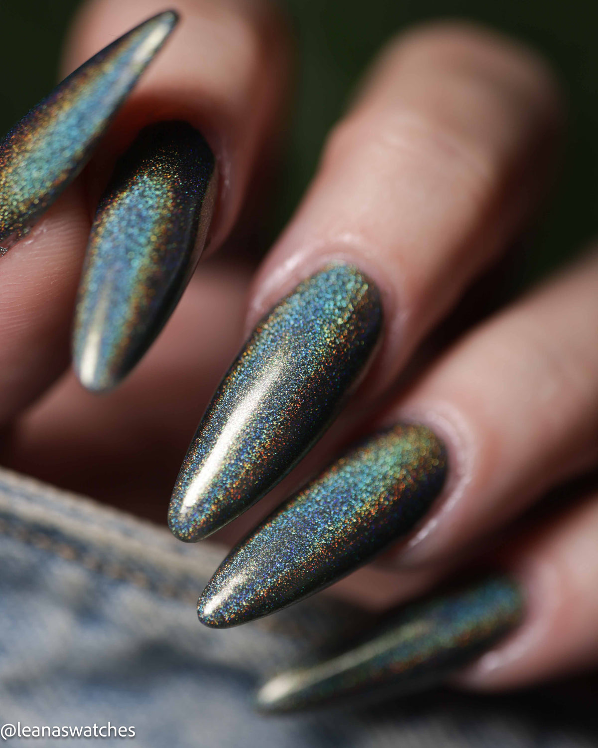 Close-up of a hand with long stiletto-shaped nails painted in a dark iridescent holographic polish that shifts between blue, green, and gold.