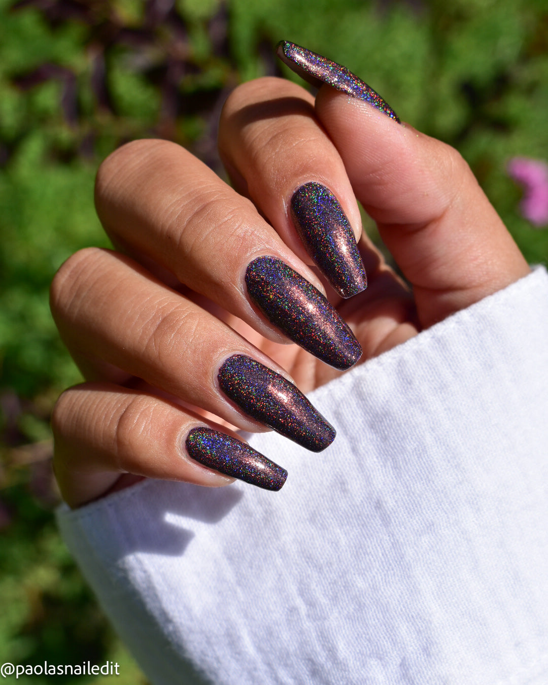 A close-up of a hand with long almond-shaped nails painted in a dark iridescent glitter nail polish, shown outdoors with a blurred green background and a white sleeve visible.