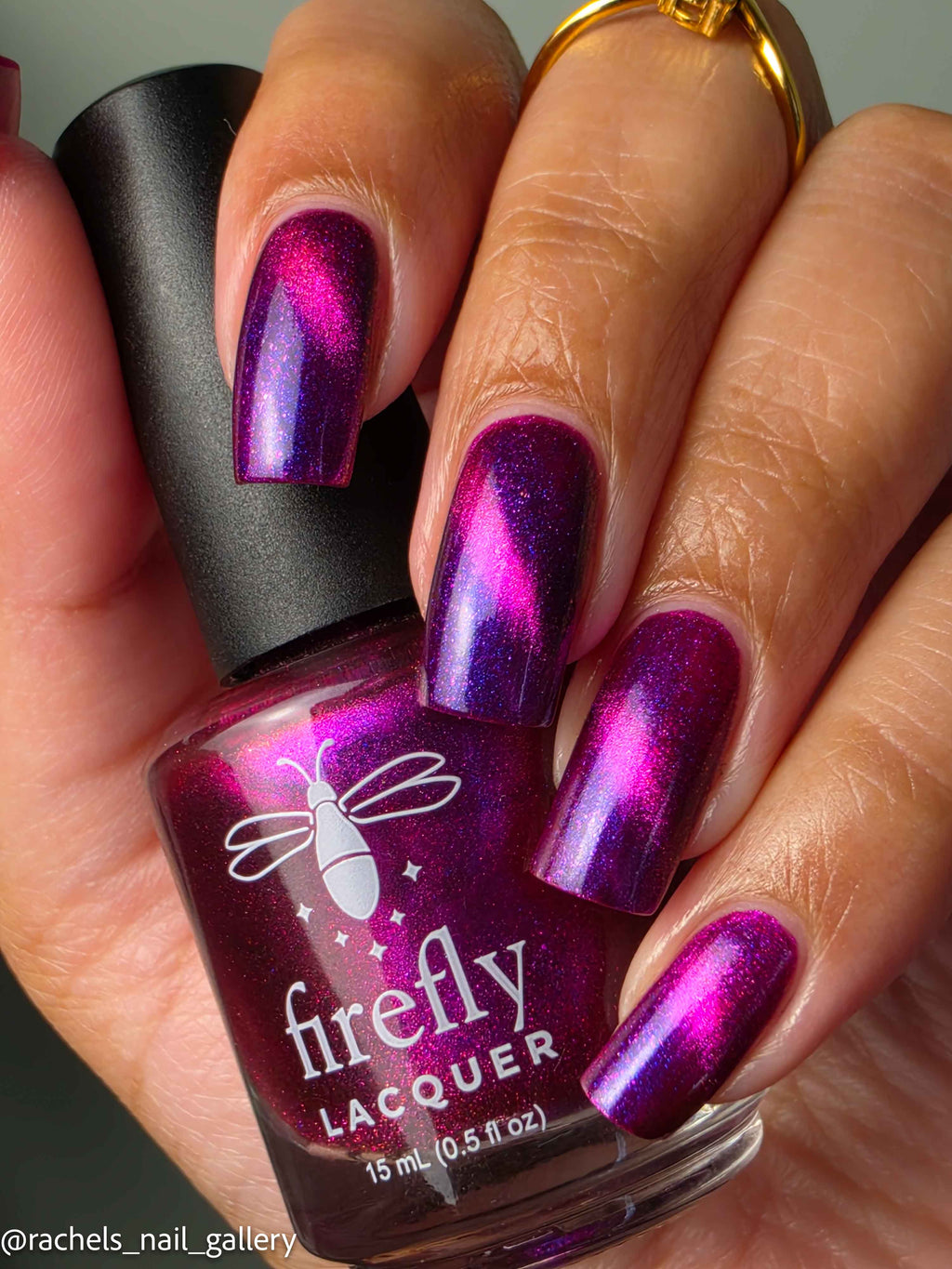 A close-up of a hand with nails painted in a metallic magenta and purple holographic lacquer, holding a bottle of Firefly Lacquer.