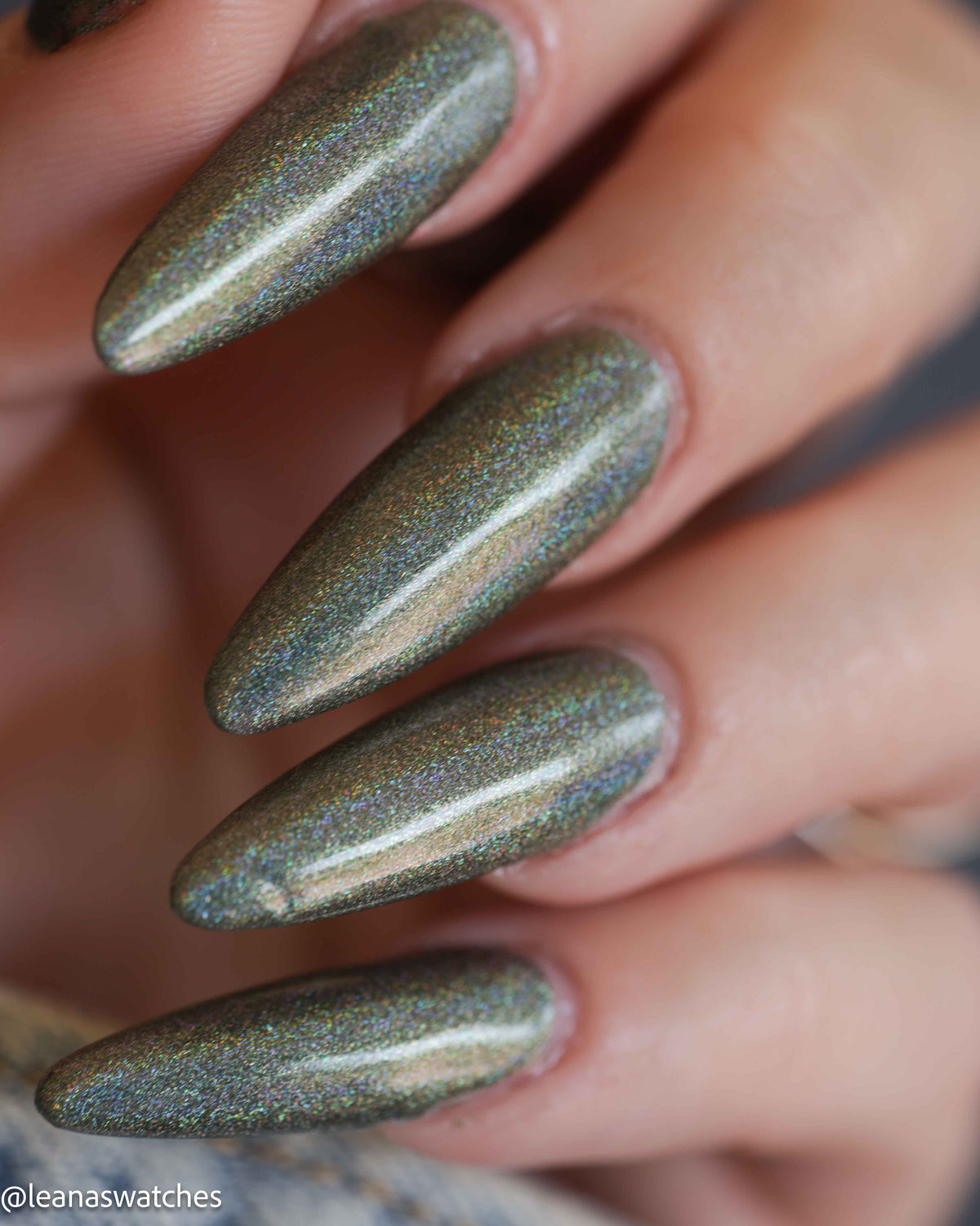 Close-up of a hand with long almond-shaped nails painted in a shimmering metallic olive green nail polish with holographic iridescent flecks.
