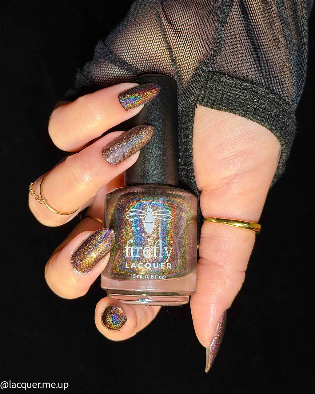 A hand holding a bottle of Firefly Lacquer nail polish against a black background. The nails are painted with a shimmering, iridescent metallic polish that shifts between bronze, gold, and purple, and the bottle itself has a black cap and a clear glass body showing the matching colored polish inside.