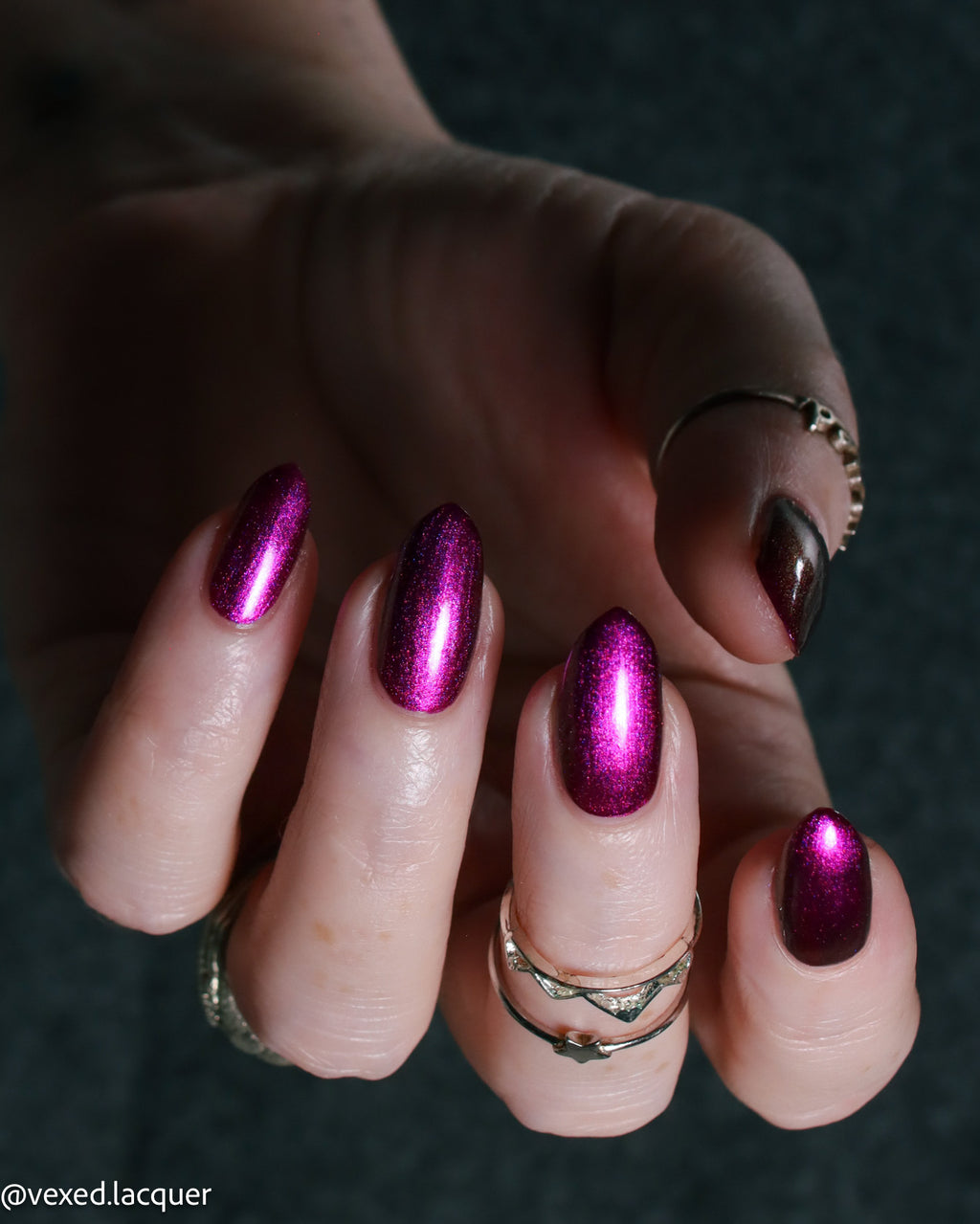 A close-up of a hand with manicured nails painted in a metallic magenta-purple color, wearing several rings against a dark textured background.