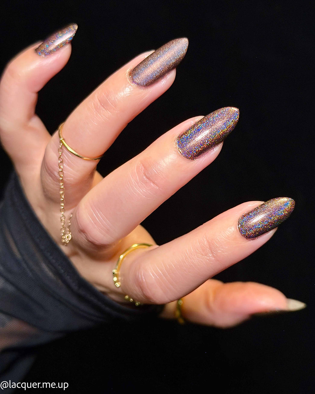 A close-up of a hand with almond-shaped nails painted in a shimmering holographic purple nail polish, accessorized with gold rings against a black background.