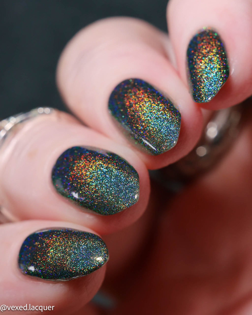 Close-up of a hand with manicured nails painted in a dark teal-blue base covered in dense iridescent holographic glitter that shifts with rainbow reflections.