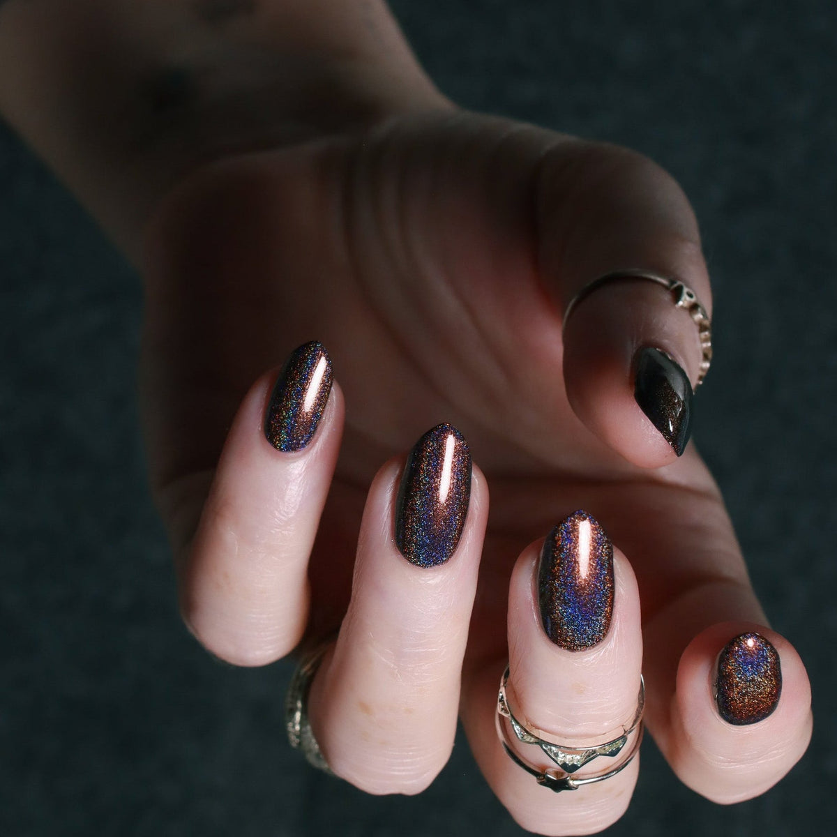 A close-up of a hand showcasing a metallic holographic nail polish with a dark base and shimmering purple, blue, and gold iridescent flecks. The hand is adorned with several rings, and the background is a dark, textured gray surface.