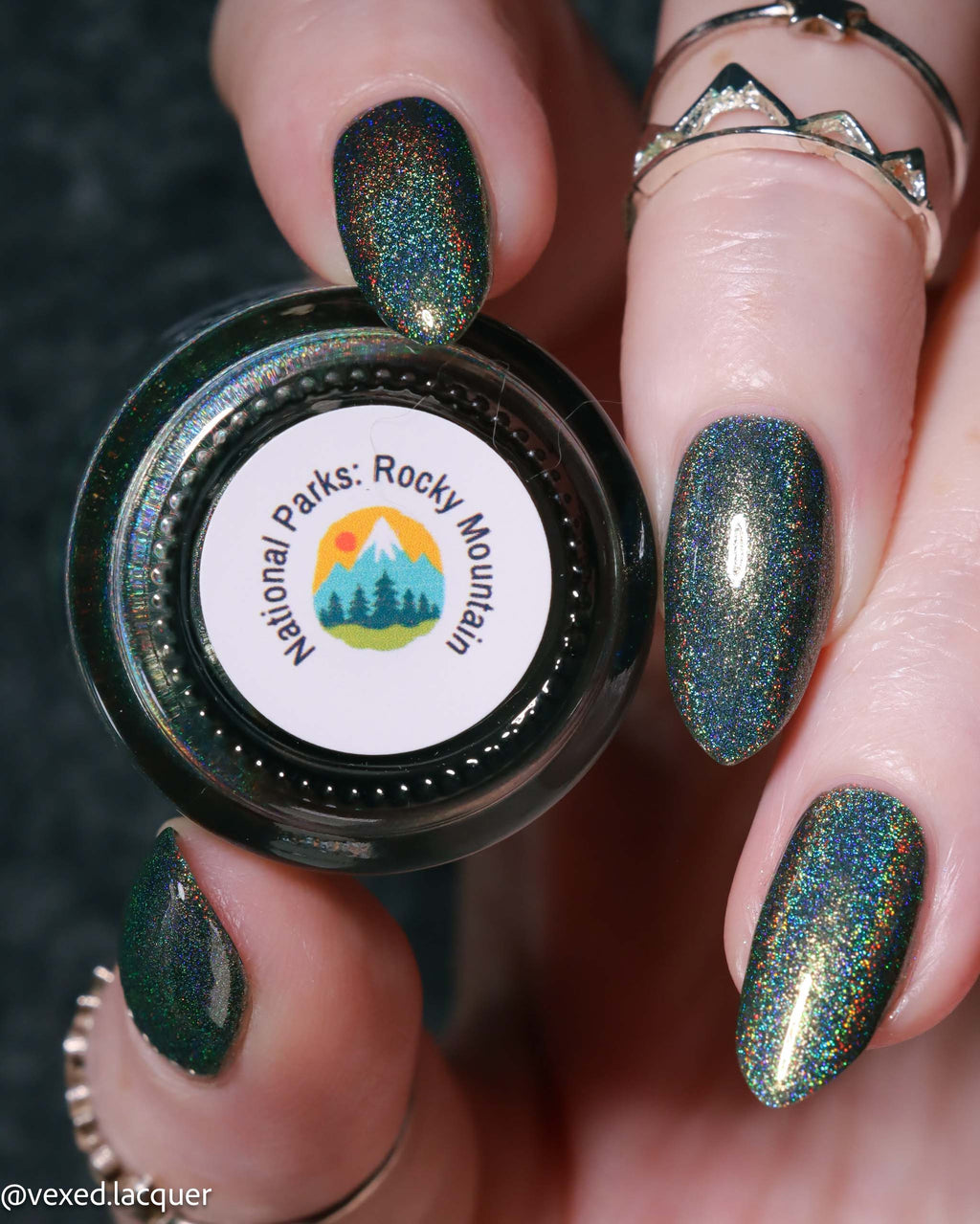A close-up of a hand with manicured nails painted in a dark green glitter nail polish with an iridescent holographic shimmer, holding a small bottle labeled 'National Parks: Rocky Mountain'.