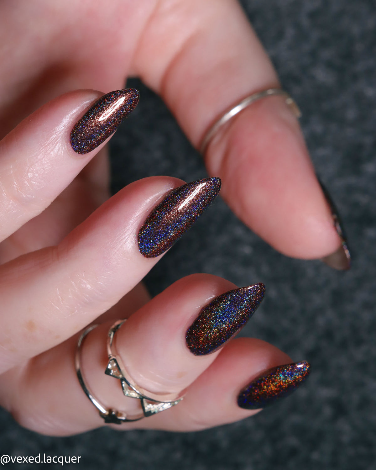 A close-up of a hand with long stiletto-shaped nails painted in a dark iridescent nail polish that shifts between bronze, blue, and orange. The hand is wearing silver rings against a dark textured background.