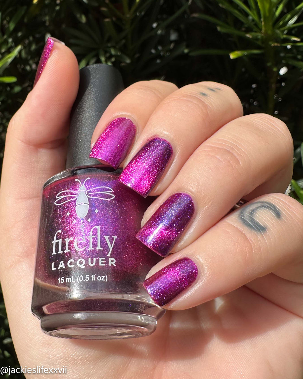 A hand holding a bottle of Firefly Lacquer nail polish in a sparkling magenta-purple shade, with the nails painted to match, set against a background of green foliage.