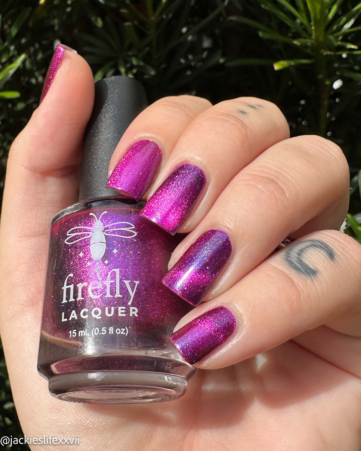 A hand holding a bottle of Firefly Lacquer nail polish in a sparkling magenta-purple shade, with the nails painted to match, set against a background of green foliage.
