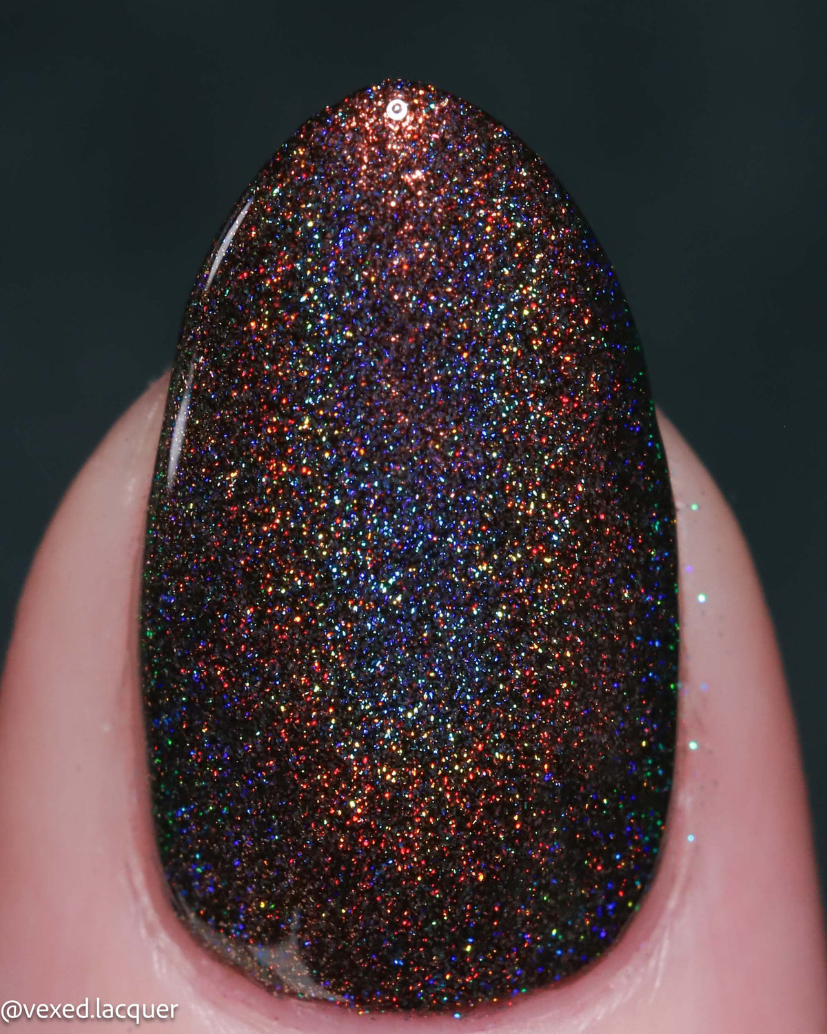 A close-up of a fingernail painted with a dark glitter nail polish featuring dense multicolor iridescent flecks over a deep brown-black base, with a dark gray background.