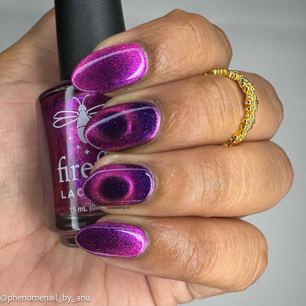 A close-up of a hand with manicured nails painted in a sparkling magenta-purple magnetic nail polish, holding a bottle of Firefly Lacquer nail polish. The hand also wears a gold ring, and the background is a plain light gray.