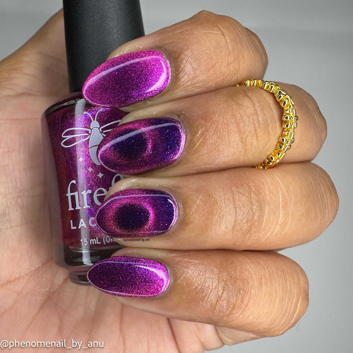 A close-up of a hand with manicured nails painted in a sparkling magenta-purple magnetic nail polish, holding a bottle of Firefly Lacquer nail polish. The hand also wears a gold ring, and the background is a plain light gray.