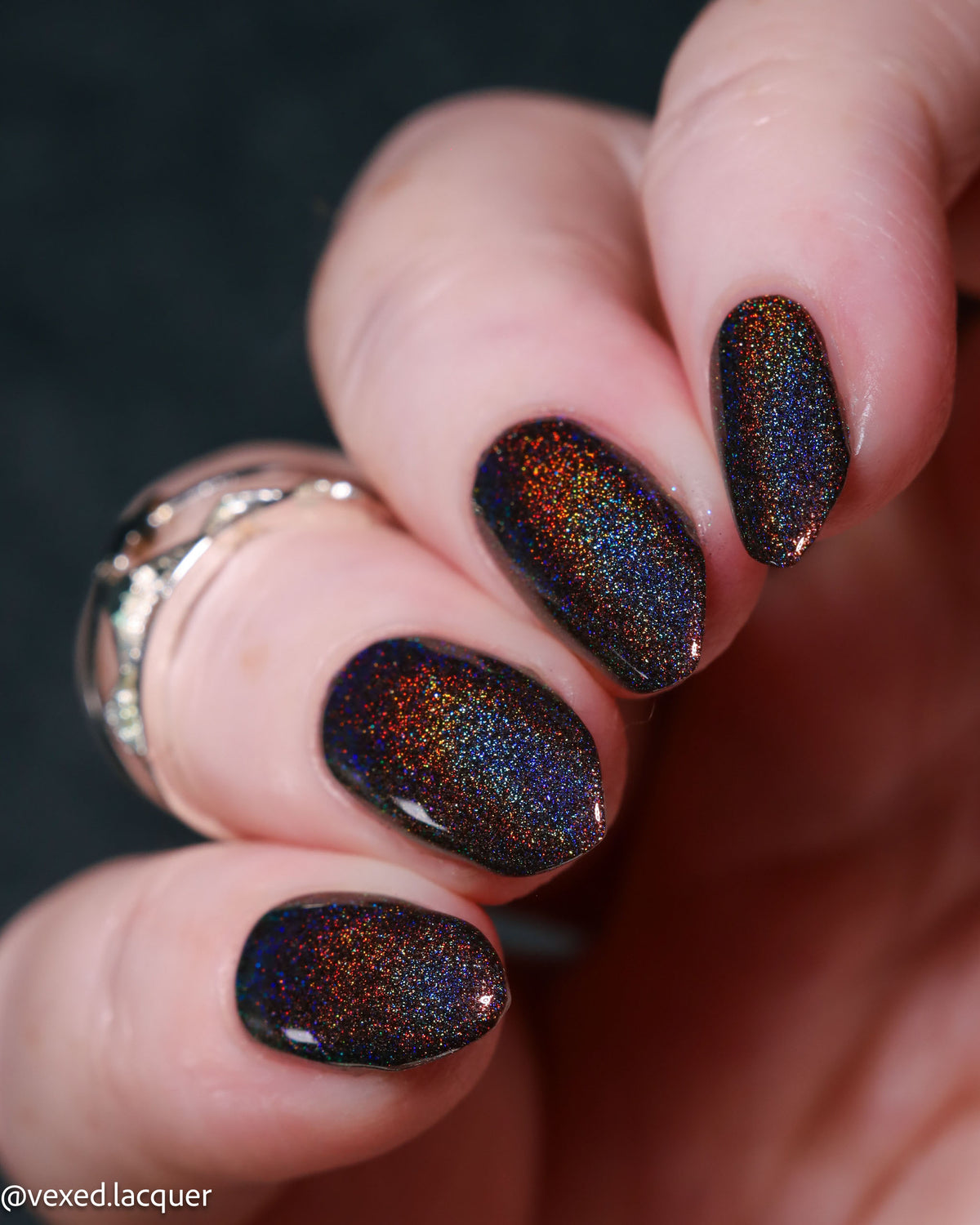 Close-up of a hand with manicured nails painted in a dark iridescent nail polish that shifts between black, blue, and copper-gold under light. A silver ring is visible on one finger, and the background is a dark, out-of-focus surface.
