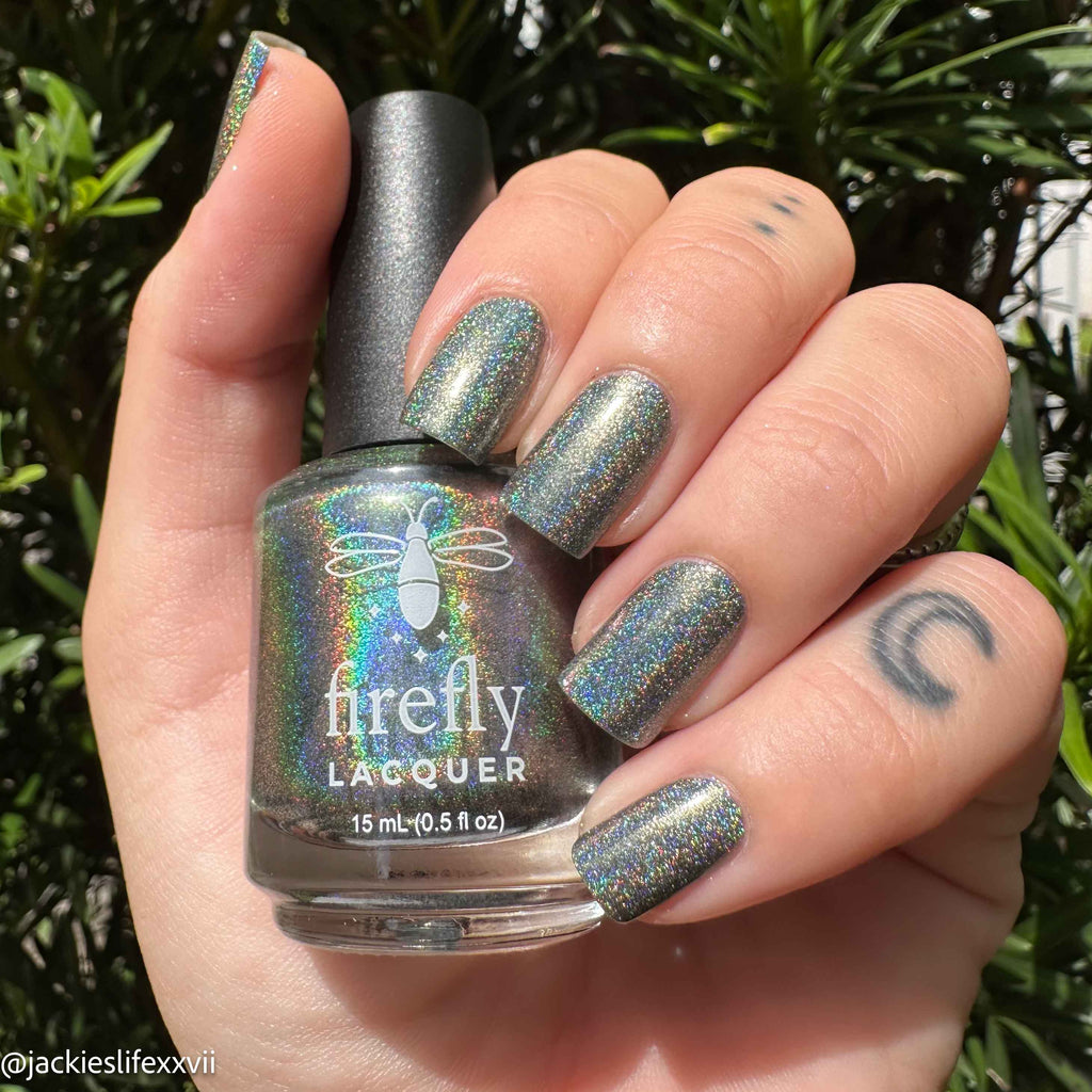 A hand holding a bottle of Firefly Lacquer nail polish with matching iridescent holographic nails painted on, set against a background of green foliage.