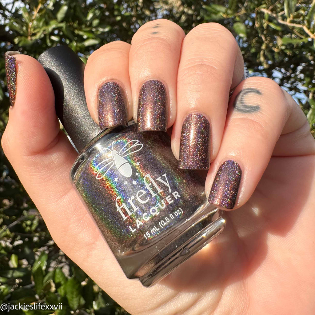 A hand holding a bottle of Freely Lacquer nail polish outdoors, with the nails painted in a dark iridescent shimmer polish that reflects purple, bronze, and green holographic tones.
