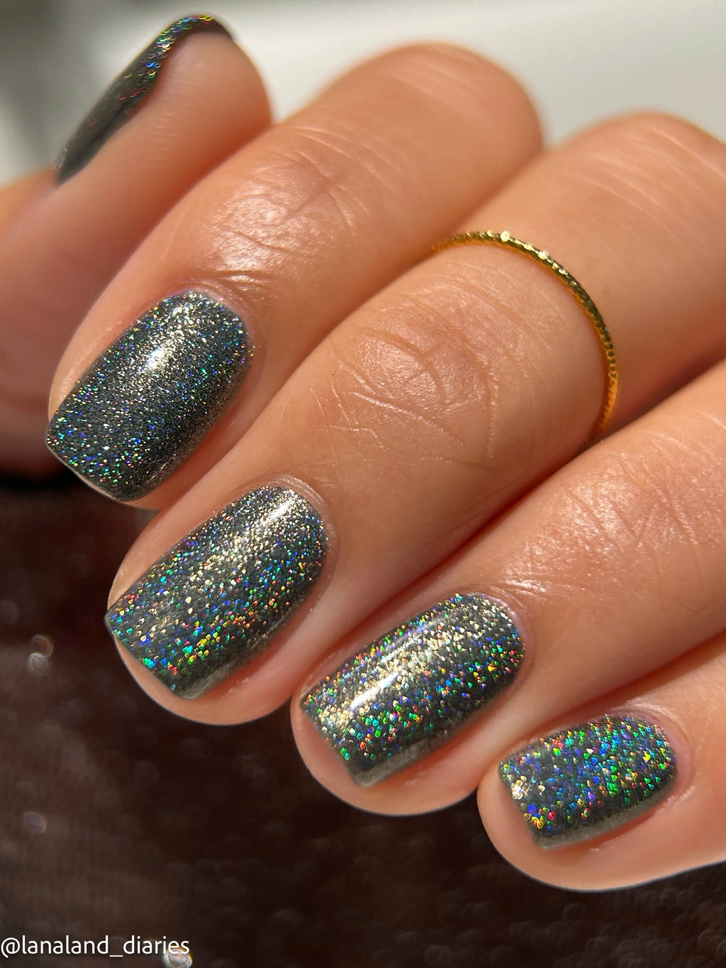 Close-up of a hand with manicured nails painted in a dark gray glitter polish that reflects holographic rainbow shimmer. A thin gold ring is worn on one finger, and the photo is cropped tightly with no visible environment beyond a dark surface.
