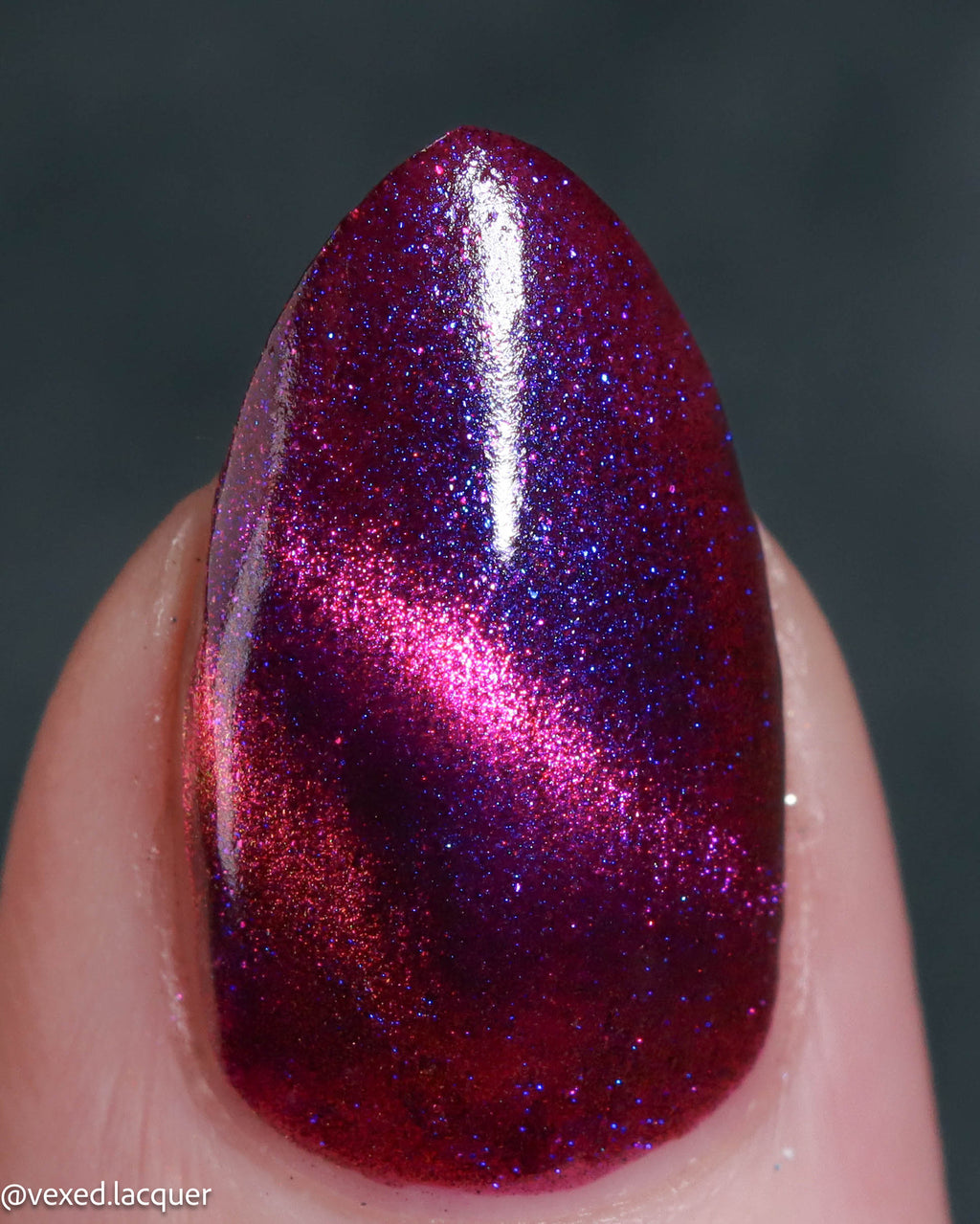 A close-up of a fingernail painted with a sparkling magnetic nail polish that shifts between deep magenta and electric blue-purple under a magnet, held against a dark gray background.