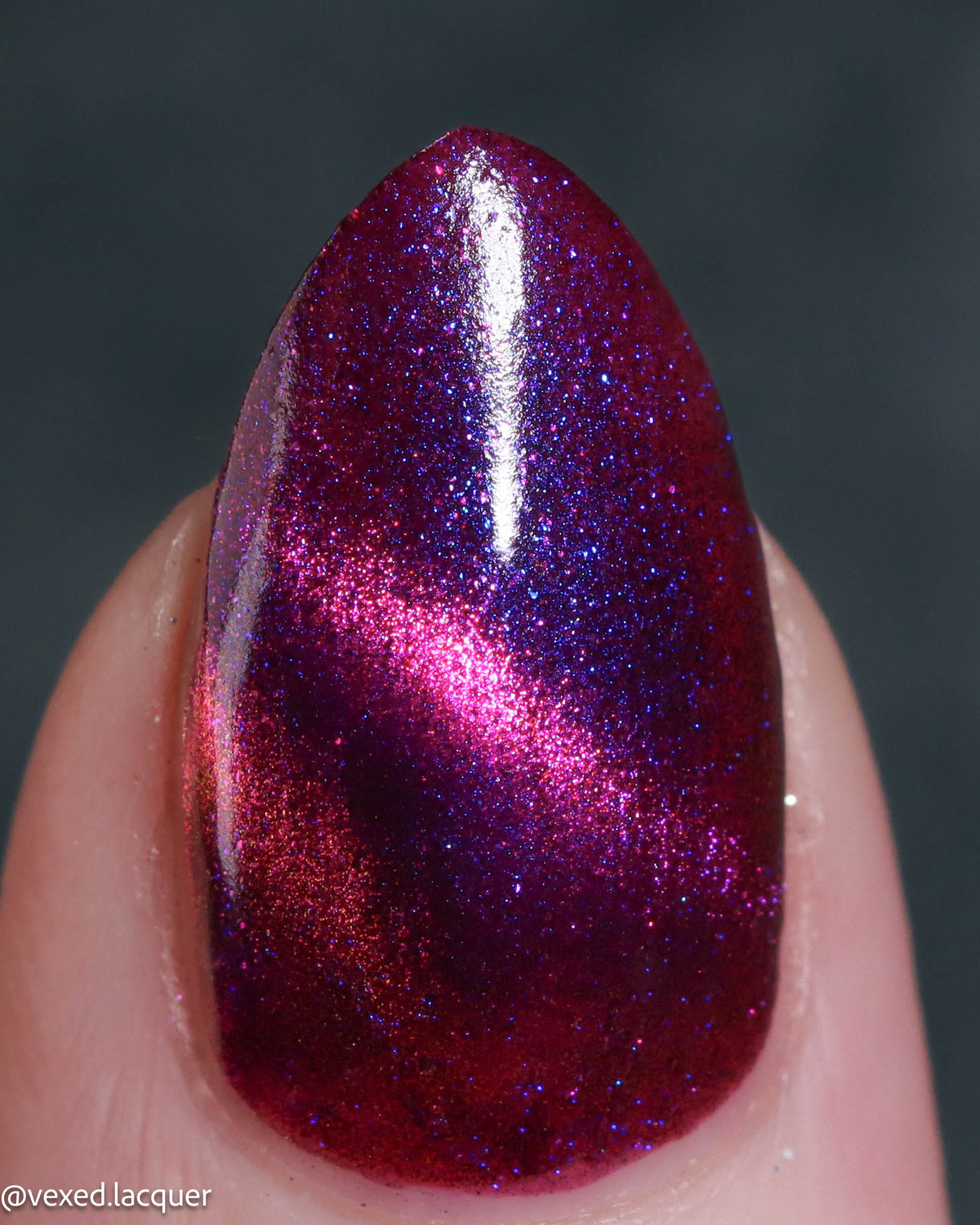 A close-up of a fingernail painted with a sparkling magnetic nail polish that shifts between deep magenta and electric blue-purple under a magnet, held against a dark gray background.