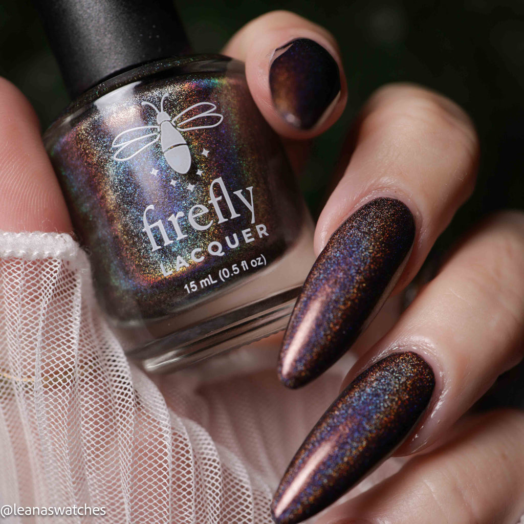 A close-up of a hand with long manicured nails painted in a dark iridescent holographic lacquer, holding a bottle labeled 'firefly LACQUER'.