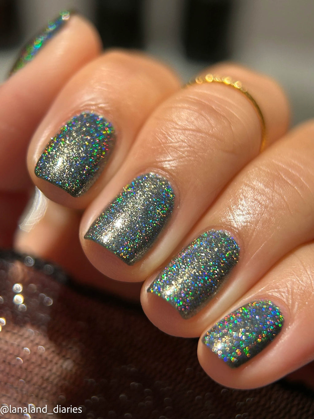 Close-up of a hand with manicured nails painted in a sparkling holographic glitter nail polish that shifts between blue, green, and silver. A gold ring is visible on one finger, and the photo is taken in warm, direct light with a blurred background.