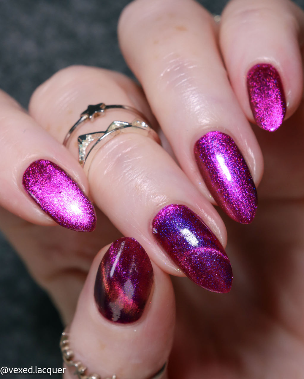 Close-up of a hand with manicured almond-shaped nails painted in a sparkling magenta-purple cat-eye nail polish, with two silver rings on the fingers.
