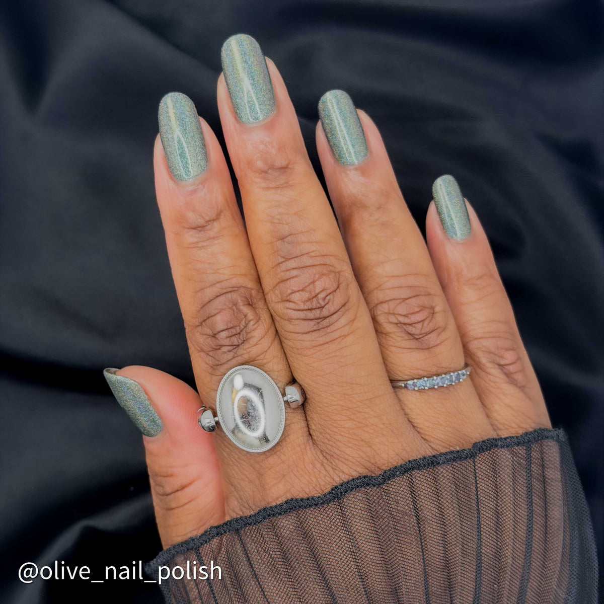 A close-up of a hand with almond-shaped nails painted in a shimmering sage green nail polish, wearing a silver ring with a large oval face and a smaller silver band with small clear stones, set against a dark fabric background.