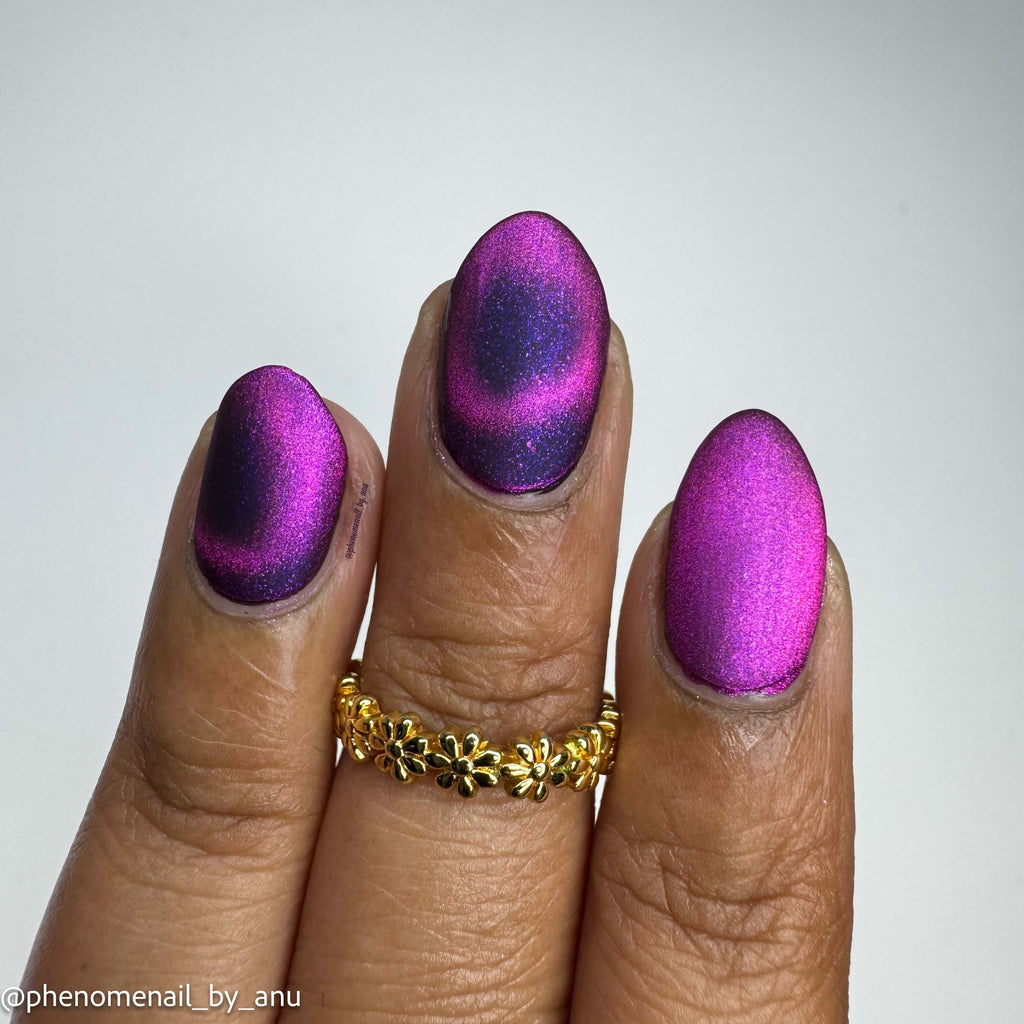 A close-up of a hand with manicured nails painted in a metallic magenta-to-purple gradient, wearing a gold ring, against a plain light gray background.
