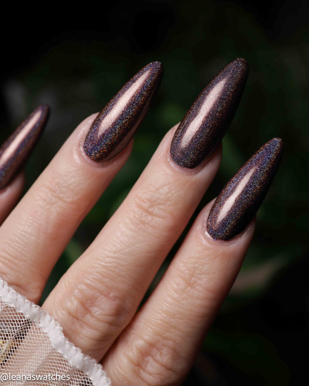 Close-up of a hand with long stiletto-shaped nails painted in a dark metallic glitter polish that shifts between deep bronze and purple with fine gold sparkle.