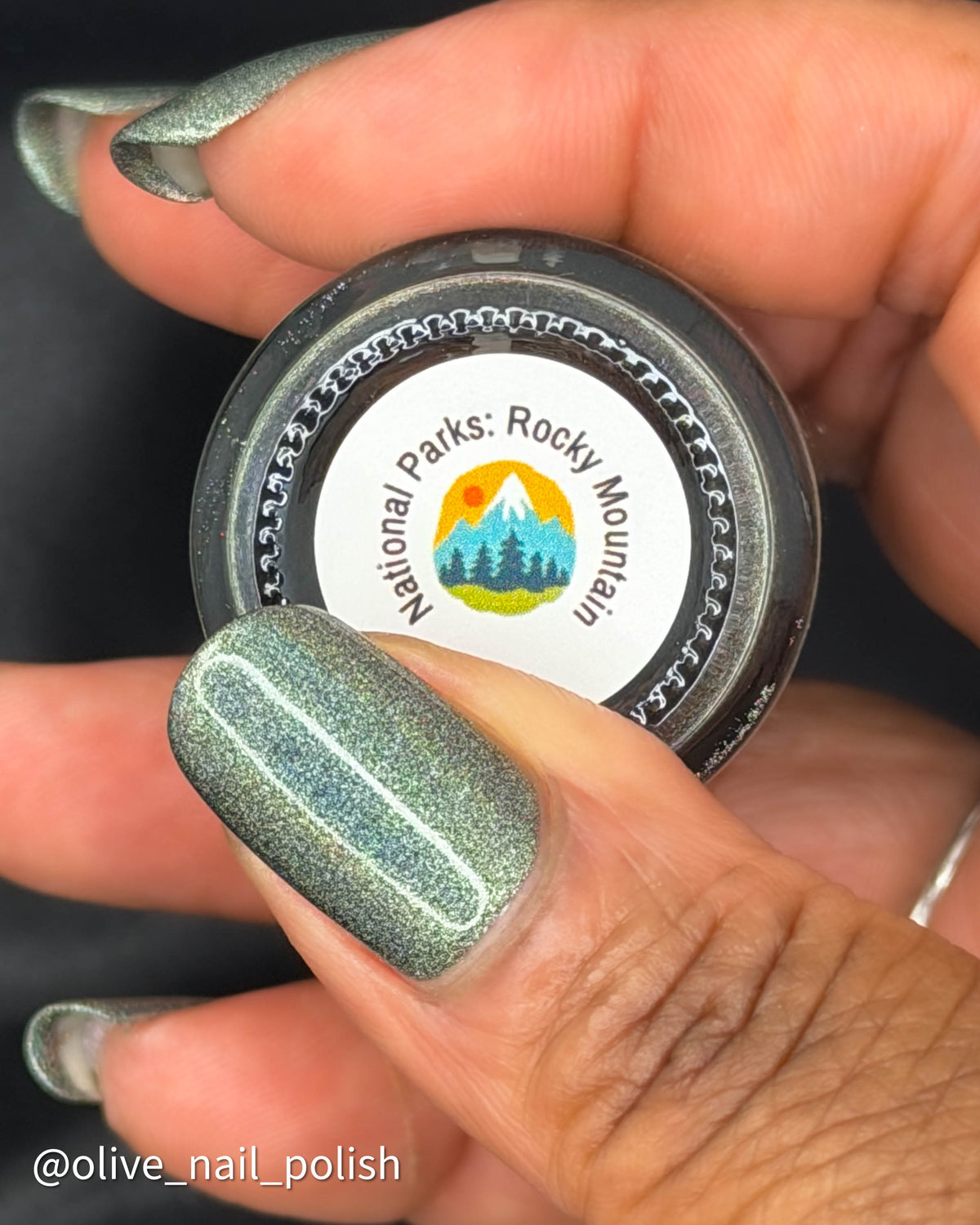 A close-up of a hand holding a small round tin of nail polish. The nail polish has a shimmering olive-sage green color with fine silver sparkle, matching the painted fingernails.