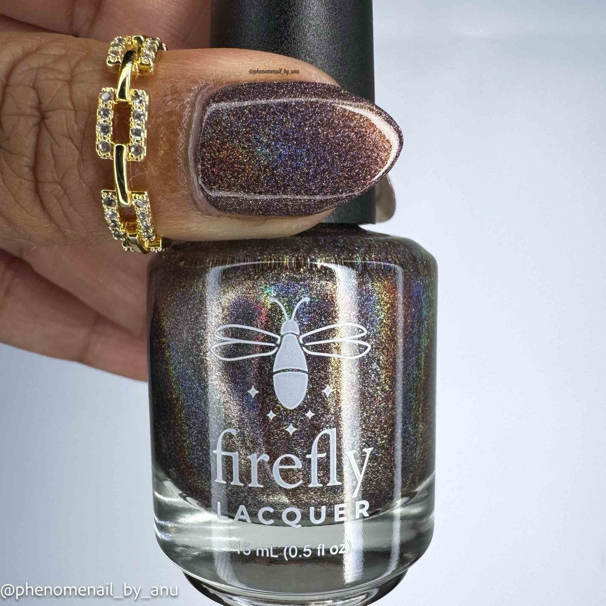 A close-up of a hand wearing a chunky gold ring and a dark iridescent holographic nail polish from Firefly Lacquer, with the bottle held in front of the hand against a light gray background.